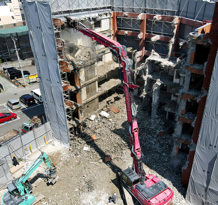 建物取り壊し（解体）作業現場。解体業者である真生工業が保有する赤いロングアーム大型重機が鉄筋コンクリート造のビル（マンション）を解体している。