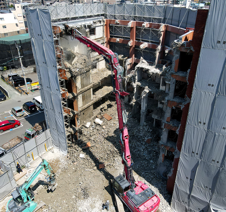 建物取り壊し（解体）作業現場。解体業者である真生工業が保有する赤いロングアーム大型重機が鉄筋コンクリート造のビル（マンション）を解体している。