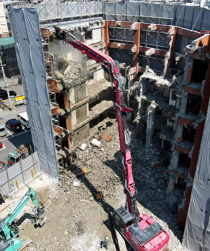 建物取り壊し（解体）作業現場。解体業者である真生工業が保有する赤いロングアーム大型重機が鉄筋コンクリート造のビル（マンション）を解体している。